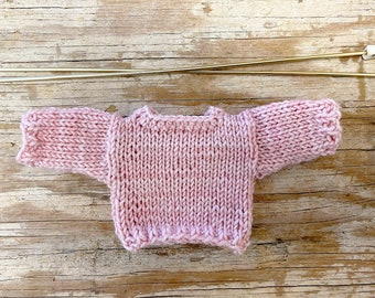 May include: A pink knitted sweater with long sleeves, laying flat on a wooden surface.