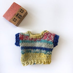 May include: A colorful striped knitted sweater with short sleeves. The sweater is made with a variety of yarn colors, including yellow, blue, green, and pink.