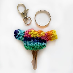 Op de afbeelding: Een sleutel met een kleurrijke, gebreide truislabels. De trui heeft regenboogkleuren, waaronder blauw, groen, geel en roze. Een zilverkleurige sleutelring en sluiting zijn ook inbegrepen.
