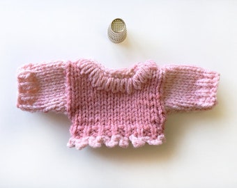 May include: A pink knitted sweater with a ruffled neckline and long sleeves.