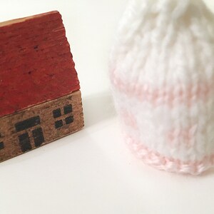 Puede incluir: Una peque&ntilde;a casa de madera con techo rojo y ventanas negras se encuentra junto a un gorro blanco tejido con una franja rosa.