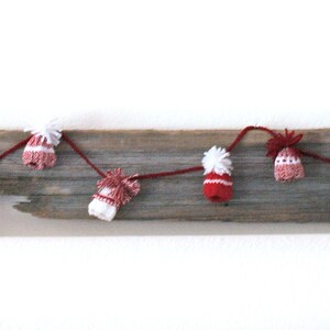 Puede incluir: Una guirnalda de gorros de punto rojos y blancos con pompones blancos encordados en una cuerda y colgados de un trozo de madera a la deriva.
