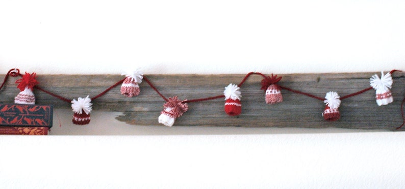Puede incluir: Una guirnalda de gorros de punto rojos y blancos con pompones blancos encordados en una cuerda y colgados de un trozo de madera a la deriva.