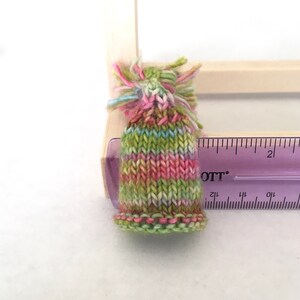 May include: A small, hand-knitted hat with a green, pink, and white striped pattern. The hat has a fluffy pom-pom on top and is about 2 inches tall.