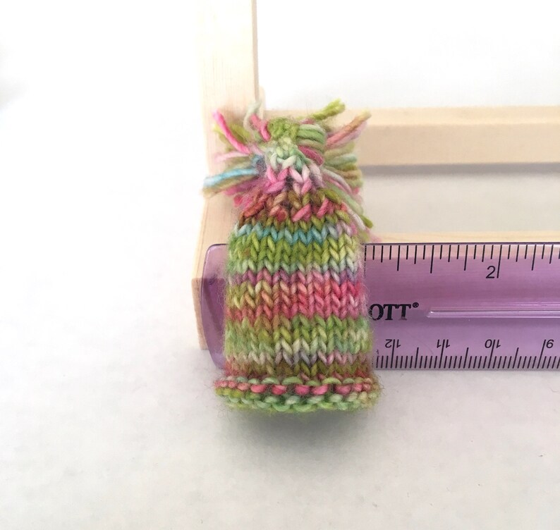 May include: A small, hand-knitted hat with a green, pink, and white striped pattern. The hat has a fluffy pom-pom on top and is about 2 inches tall.