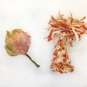 Puede incluir: Un pequeño gorro tejido a mano con un patrón de rayas blancas y naranjas. El gorro tiene un pompón esponjoso en la parte superior y está sentado sobre una superficie blanca junto a una hoja seca.