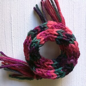 May include: A knitted scarf with pink, green, and purple yarn. The scarf has fringe on both ends.