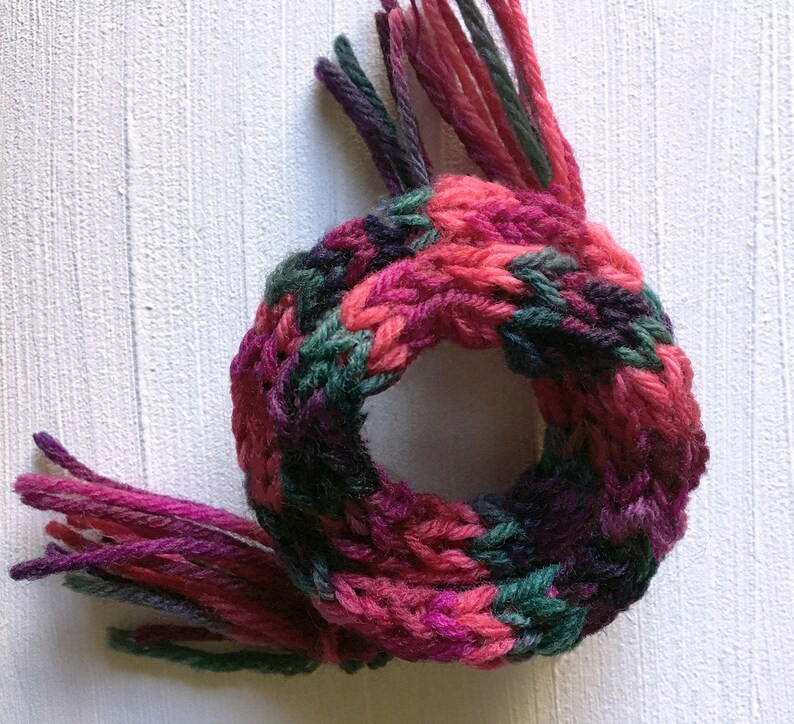 May include: A knitted scarf with pink, green, and purple yarn. The scarf has fringe on both ends.
