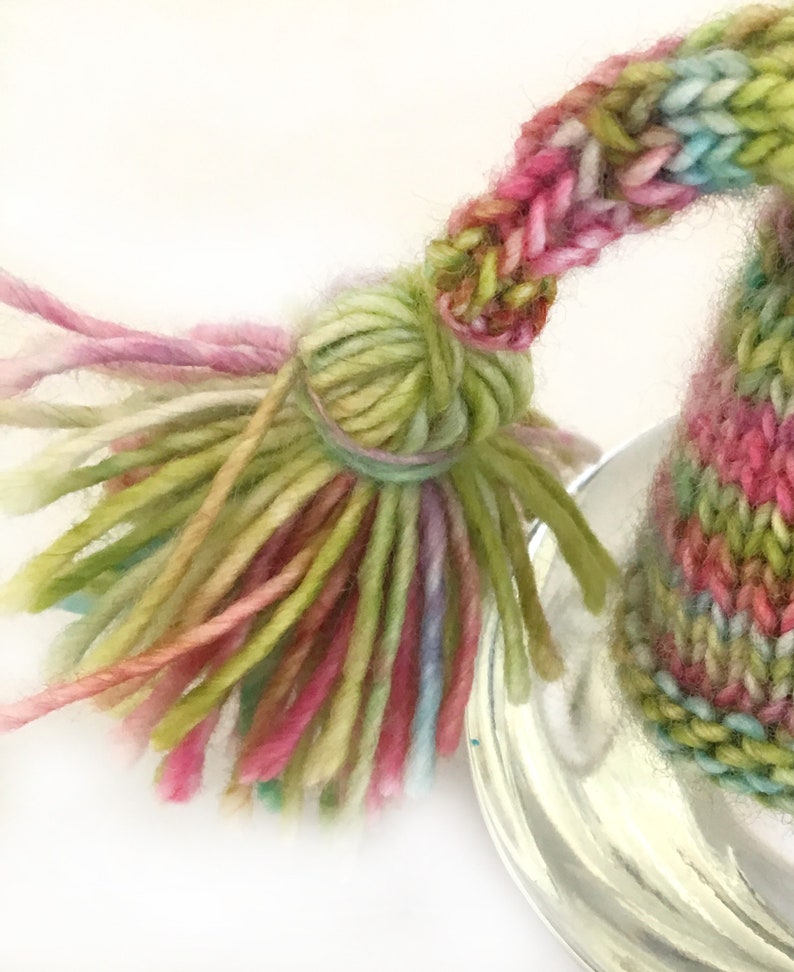 May include: A colorful knitted hat with a long tassel. The hat is made with a variety of shades of pink, green, and blue yarn.