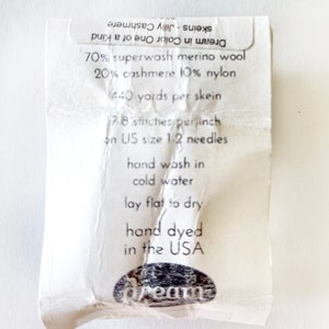 May include: A white label with black text detailing the yarn's composition, yardage, needle size, and care instructions. The label reads "70% superwash merino wool 20% cashmere 10% nylon 1440 yards per skein 17.8 stitches per inch on US size 1-2 needles hand wash in cold water lay flat to dry hand dyed in the USA Dream in Color One of a Kind skeins - Jilly Cashmere".