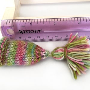 May include: Two colorful knitted yarn tassels, one with a pointed tip and the other with a rounded tip, measured against a ruler. The tassels are approximately 2 inches long and feature a variety of colors, including pink, green, and white.