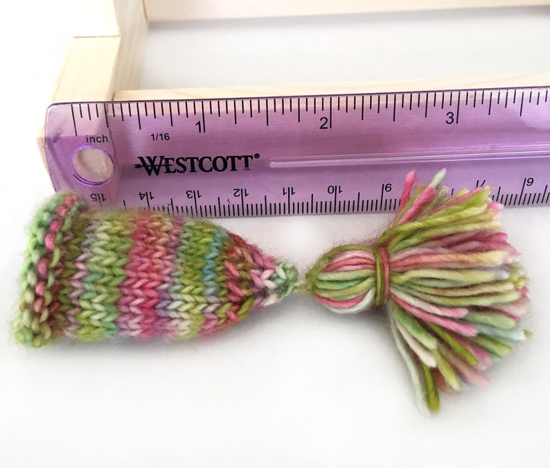 May include: Two colorful knitted yarn tassels, one with a pointed tip and the other with a rounded tip, measured against a ruler. The tassels are approximately 2 inches long and feature a variety of colors, including pink, green, and white.