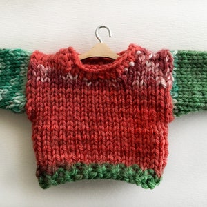 May include: A red, green, and white knitted sweater with a wooden hanger. The sweater has a green trim around the neck and bottom.