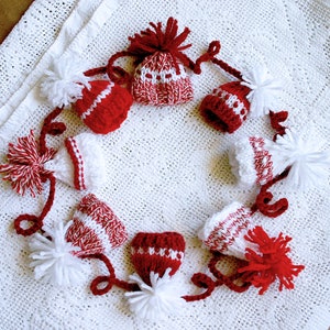 Puede incluir: Una guirnalda de gorros de punto rojos y blancos con pompones blancos. Los gorros est&aacute;n conectados por hilo rojo y blanco.