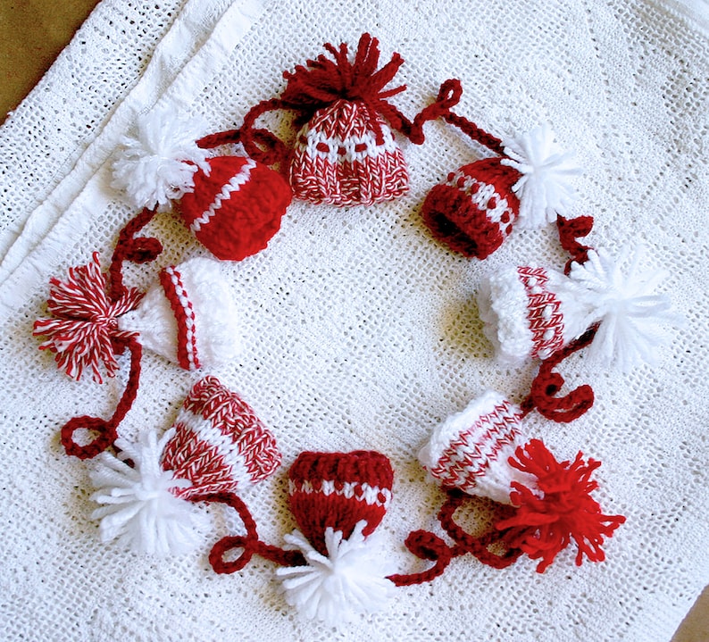 Puede incluir: Una guirnalda de gorros de punto rojos y blancos con pompones blancos. Los gorros est&aacute;n conectados por hilo rojo y blanco.