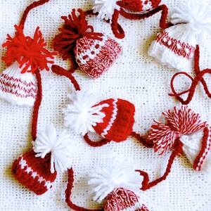 Puede incluir: Una guirnalda de gorros de punto rojos y blancos con pompones blancos. Los gorros est&aacute;n conectados por una cuerda blanca.