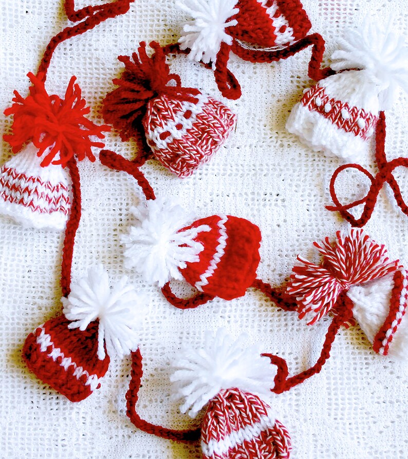 Puede incluir: Una guirnalda de gorros de punto rojos y blancos con pompones blancos. Los gorros est&aacute;n conectados por una cuerda blanca.