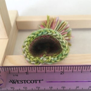 May include: A close-up of a green, pink, and yellow crocheted hat with a pom-pom on top. The hat is measured against a ruler, showing it is about 2 inches in diameter.