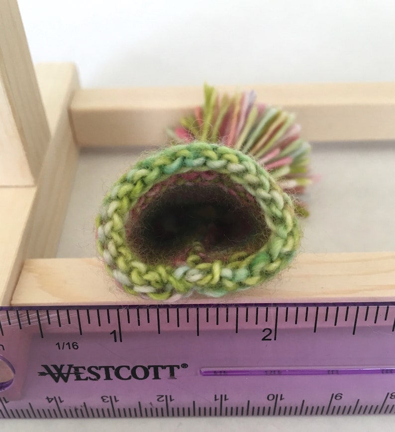 May include: A close-up of a green, pink, and yellow crocheted hat with a pom-pom on top. The hat is measured against a ruler, showing it is about 2 inches in diameter.