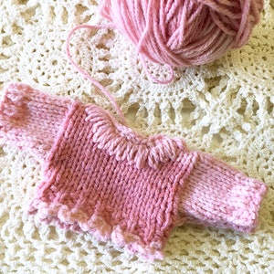 May include: A pink knitted sweater with a ruffled neckline and sleeves. The sweater is lying on a white crocheted surface.