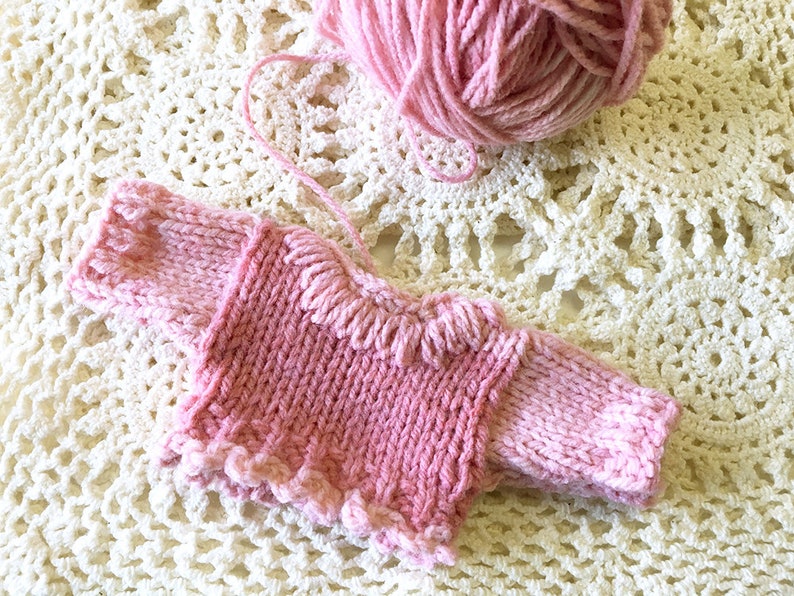 May include: A pink knitted sweater with a ruffled neckline and sleeves. The sweater is lying on a white crocheted surface.