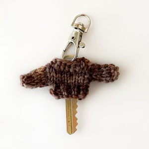 May include: A brown knitted sweater key chain with a silver clasp. The key chain is made of brown and tan yarn and is shaped like a miniature sweater. The key is inserted through the sweater.