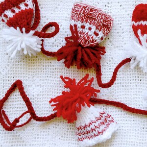Puede incluir: Tres gorros de punto rojo y blanco con pompones blancos en la parte superior. Los gorros est&aacute;n conectados por un hilo de lana rojo.