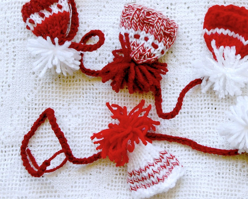 Puede incluir: Tres gorros de punto rojo y blanco con pompones blancos en la parte superior. Los gorros est&aacute;n conectados por un hilo de lana rojo.