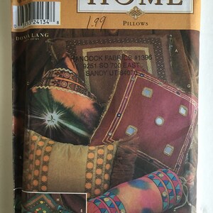 May include: A Simplicity Home sewing pattern for pillows. The pattern shows a variety of pillows with different designs and colors, including a brown pillow with gold trim, a red pillow with gold trim, a blue and green pillow with gold trim, and a long, cylindrical pillow with a colorful design. The pattern also includes the text "Simplicity Home" and "Pillows".