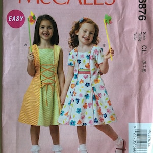 May include: McCall's sewing pattern M6876 for girls' dresses. The pattern features two girls wearing different dresses. One dress is yellow and green with a lace-up front, and the other is white with a floral print. The pattern is labeled "Easy."