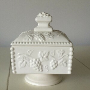 May include: A white ceramic trinket box with a lid, featuring a raised grape and leaf design. The box is square with rounded edges and sits on a pedestal base. The lid has a small, decorative handle. The box is approximately 6 inches tall.