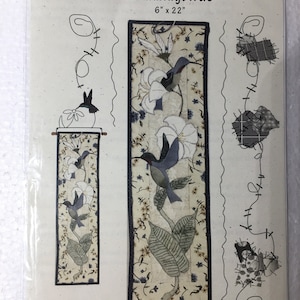 May include: A package for a fabric craft project titled "Hummingbirds" by Patch Abilities, Inc. The design features hummingbirds, white flowers, and green leaves on a beige background. The dimensions are 6" x 22".