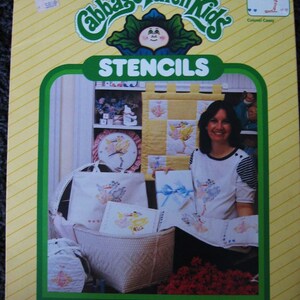 May include: A package of Cabbage Patch Kids stencils featuring a yellow and white quilt with a stork design. The package includes two pre-cut 8 1/2 by 11 inch stencil sheets. The package is yellow with green and white stripes. The text "Quality Stencils of Mylar for Home & Fashion Decor" is printed on the bottom of the package.