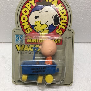 May include: A vintage Snoopy Handfuls Mini Die Cast Wagon toy featuring Charlie Brown in a blue wagon with yellow wheels. The packaging includes Snoopy and Woodstock graphics, with the text "CHARLIE BROWN" on the wagon.