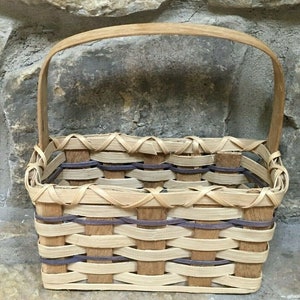 May include: A rectangular woven basket with a wooden handle. The basket is made of light brown and dark brown woven strips, with a dark purple accent. The handle is made of light brown wood.