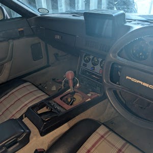 Puede incluir: Vista interior de un coche Porsche clásico. El salpicadero es oscuro con indicadores y una consola central. Los asientos tienen un estampado de cuadros. El volante tiene el logotipo de Porsche. El interior del coche muestra signos de desgaste.