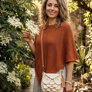 Peut inclure: Une femme porte un poncho rouille et une robe beige, tenant un petit sac à main en crochet blanc en forme de hibou avec un petit sac assorti. Les sacs ont des lanières en cuir marron. L'arrière-plan présente une verdure luxuriante et des fleurs blanches.