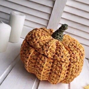 以下が含まれることがあります： オレンジと黄色の手作りの質感のあるカボチャの装飾。緑色の茎が付いています。織り込まれた素材で作られたカボチャは、白い表面に置かれ、居心地の良い秋のテーマを連想させます。