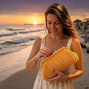 May include: A mustard yellow, rectangular crochet clutch with a rounded top. The clutch is held by a person wearing a cream-colored dress with a beach sunset in the background. The clutch has a ribbed texture.