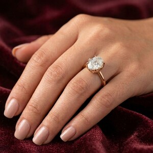 Peut inclure: Une bague ornée d'une grande pierre précieuse transparente de taille ovale, montée sur un anneau en or rose poli. La pierre est maintenue par un sertissage à griffes multiples qui l'élève au-dessus de l'anneau, révélant une surface facettée.