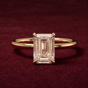 Pode incluir: Um anel de ouro com uma grande pedra preciosa transparente de corte esmeralda retangular, fixada numa cravação de quatro garras. A banda é lisa e polida, apresentando um design clássico com um acabamento brilhante que reflete a luz.