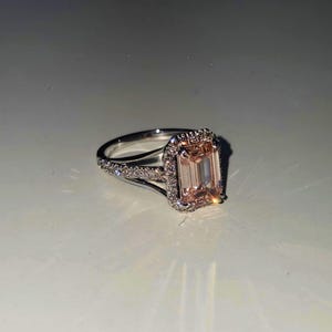 May include: A silver-toned ring featuring a large, rectangular, peach-colored center stone in an emerald cut. The center stone is surrounded by a halo of small, clear, round-cut stones, with additional small stones set along the split-style band.