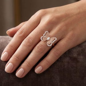 May include: Rose gold butterfly ring with a central diamond and smaller surrounding stones. The ring's butterfly design features openwork wings and is set on a delicate band. The ring is shown on a hand with a neutral nail polish.