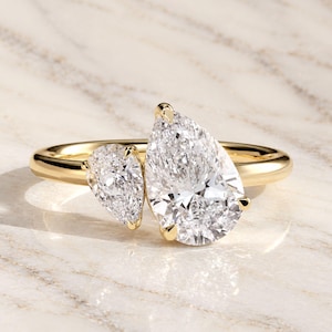 Pode incluir: Um anel de ouro amarelo com duas pedras preciosas transparentes em lapidação pera num estilo bypass. A pedra maior em forma de pera está posicionada ao lado de uma menor, ambas fixadas com garras de ouro numa banda de ouro polido.