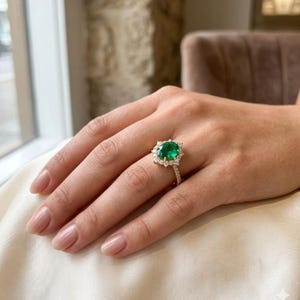 May include: A gold ring featuring a large oval-cut green gemstone at the center, surrounded by a halo of small round white stones. The band is thin and also set with small white stones along the visible portion of the shank, creating a bright sparkle.