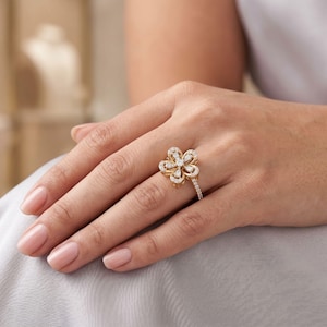Könnte beinhalten: Ein goldener Ring mit einem blumenförmigen Mittelstück, das mit mehreren kleinen, klaren, funkelnden Steinen besetzt ist. Die Ringschiene ist schmal und ebenfalls mit einer Reihe winziger, klarer Steine im Brillantschliff verziert.