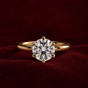 Op de afbeelding: Een gouden ring met een grote, ronde, briljant geslepen heldere edelsteen die op zijn plaats wordt gehouden door een zetting met zes pootjes. De ringband is glad, gepolijst en rond, met een klassiek ontwerp en een glanzende metalen afwerking.