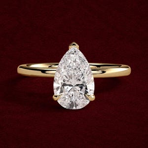 Pode incluir: Um anel de ouro com uma única pedra preciosa transparente lapidada em forma de pera e fixada por garras. A banda é lisa e polida, apresentando um design simples que destaca a silhueta alongada e em forma de gota da pedra central.