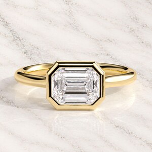 Pode incluir: Um anel de ouro com uma pedra preciosa transparente de corte esmeralda retangular, montada num engaste liso e polido. A banda é de ouro amarelo maciço com um perfil arredondado, apresentando uma superfície limpa, reflexiva e simétrica.