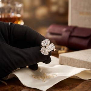 May include: A gold ring with three large diamonds in oval, emerald, and pear cuts. The ring is held by a gloved hand over a white cloth with gold embroidery. The background is blurred, suggesting a luxury setting.